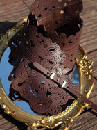 The Vintage Floral Cutout Waist Belt by Social Queen, featuring intricate laser-cut details and a scalloped edge in top grain leather, is showcased on an ornate gold-framed mirror atop wooden planks.