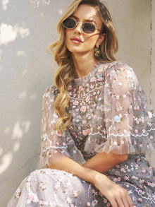 A woman with long wavy hair wears Social Queen’s Vintage Gold-Trimmed Black Sunglasses and a sheer floral dress, sitting against a light wall in natural sunlight.