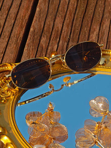 Social Queen’s Vintage Gold-Trimmed Black Sunglasses with dark lenses rest on a gold mirror atop wood, reflecting metallic floral accents.