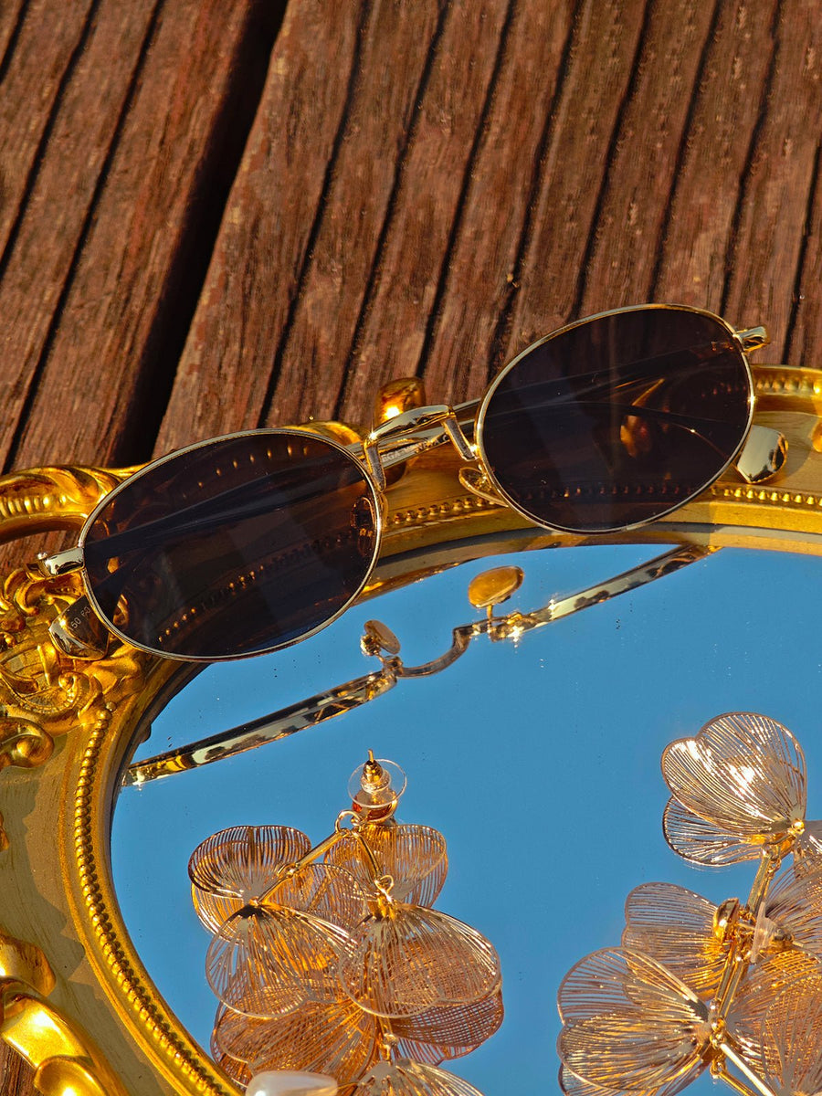 The Social Queen Vintage Gold-Trimmed Black Sunglasses with high-clarity dark lenses rest on a gold-framed mirror on wooden boards, reflecting metallic floral ornaments.