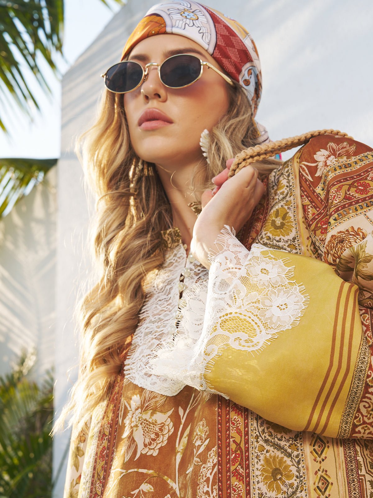 A woman stands outdoors among palm leaves, wearing Social Queen's Vintage Gold-Trimmed Black Sunglasses, a patterned headscarf, and a floral lace-trimmed dress. Sunlight streams in, highlighting her vintage-inspired look.