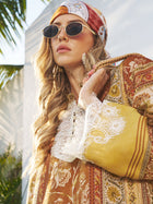 A woman stands outdoors among palm leaves, wearing Social Queen's Vintage Gold-Trimmed Black Sunglasses, a patterned headscarf, and a floral lace-trimmed dress. Sunlight streams in, highlighting her vintage-inspired look.