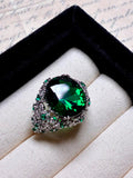 The Social Queen Vintage Green Crystal Ring features an oval-cut green gemstone with smaller green and clear stones, a white gold plated adjustable open band, and comes elegantly displayed in a jewelry box.