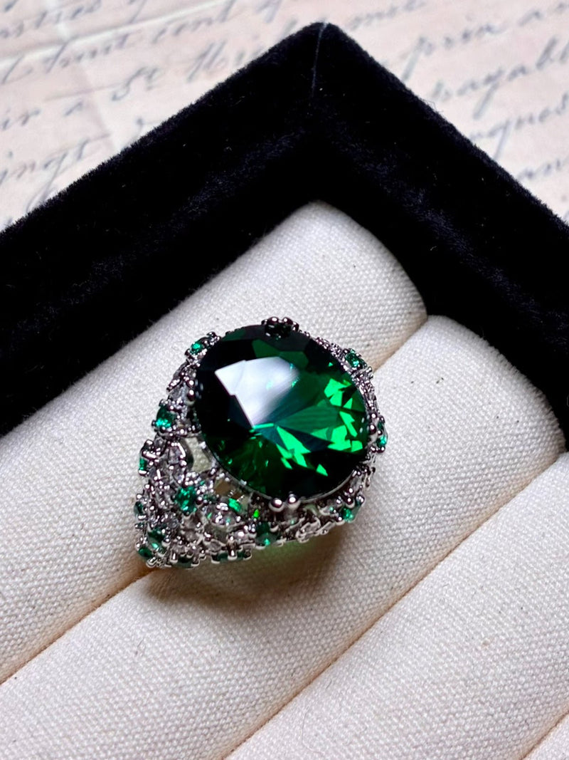 The Social Queen Vintage Green Crystal Ring features a large oval green gemstone with smaller green and clear stones, set on a white gold-plated adjustable open band and presented in a black and cream ring box.