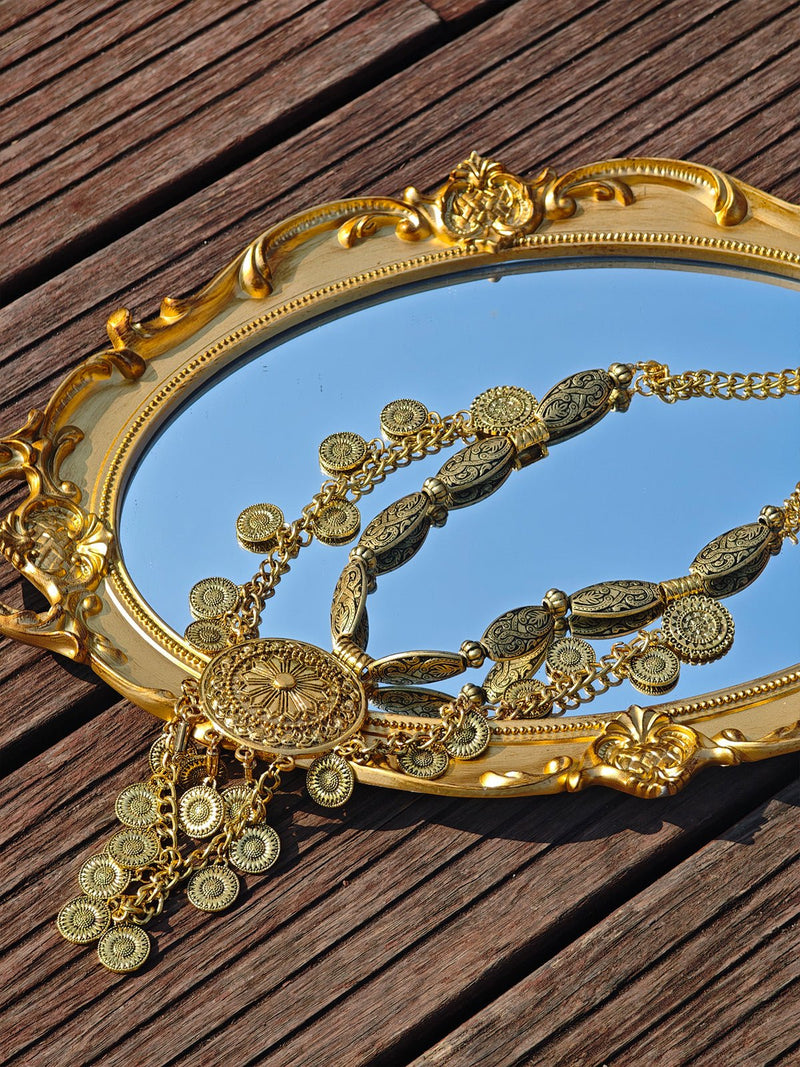 The Vintage Sunflower Necklace by Social Queen, featuring intricate sunflower motifs and gold plating, is elegantly displayed on an ornate oval mirror with a gold frame atop a wooden surface.