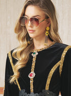 A woman in Social Queen’s Vintage Sunglasses with Gold Frame, a black and gold embellished dress, and statement earrings stands against a neutral background.