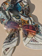 A pair of Social Queen Vintage Sunglasses with a gold frame rests on a multicolored patterned scarf atop a light surface.