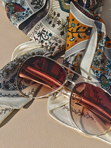 The Social Queen Vintage Sunglasses with a gold frame and brown-tinted lenses are displayed on a floral-patterned scarf atop a beige surface.