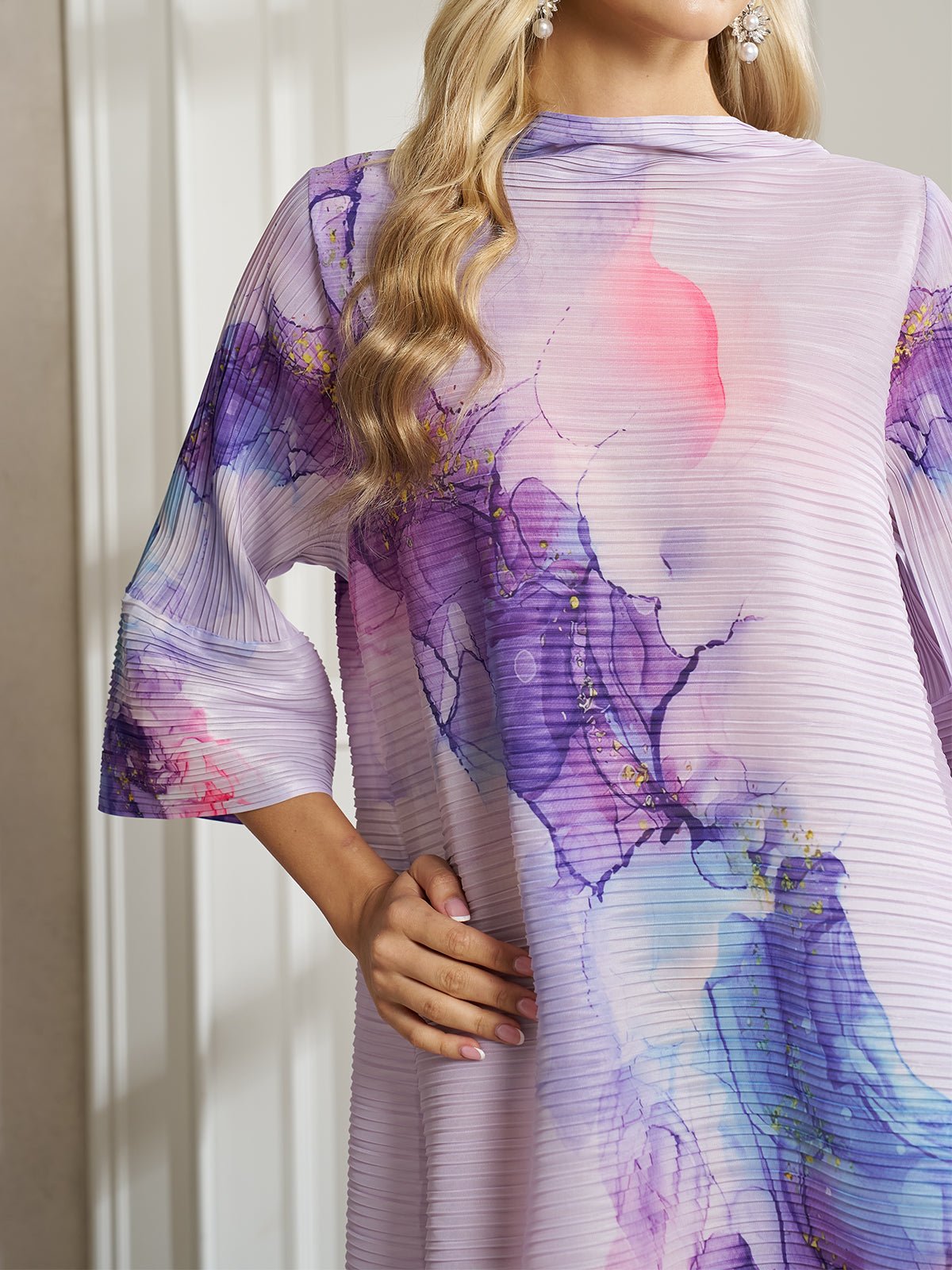 A woman with wavy blonde hair stands confidently with one hand on her hip, showcasing effortless style in the Social Queen Watercolor Print Plissé Dress in pastel pink, blue, and purple hues.