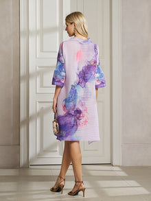 A woman stands indoors facing a white door, wearing the Social Queen Watercolor Print Plissé Dress in purple and pink patterns and holding a small handbag.