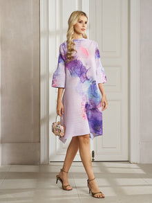 A woman stands indoors wearing the Social Queen Watercolor Print Plissé Dress, paired with a small floral handbag and heeled sandals.