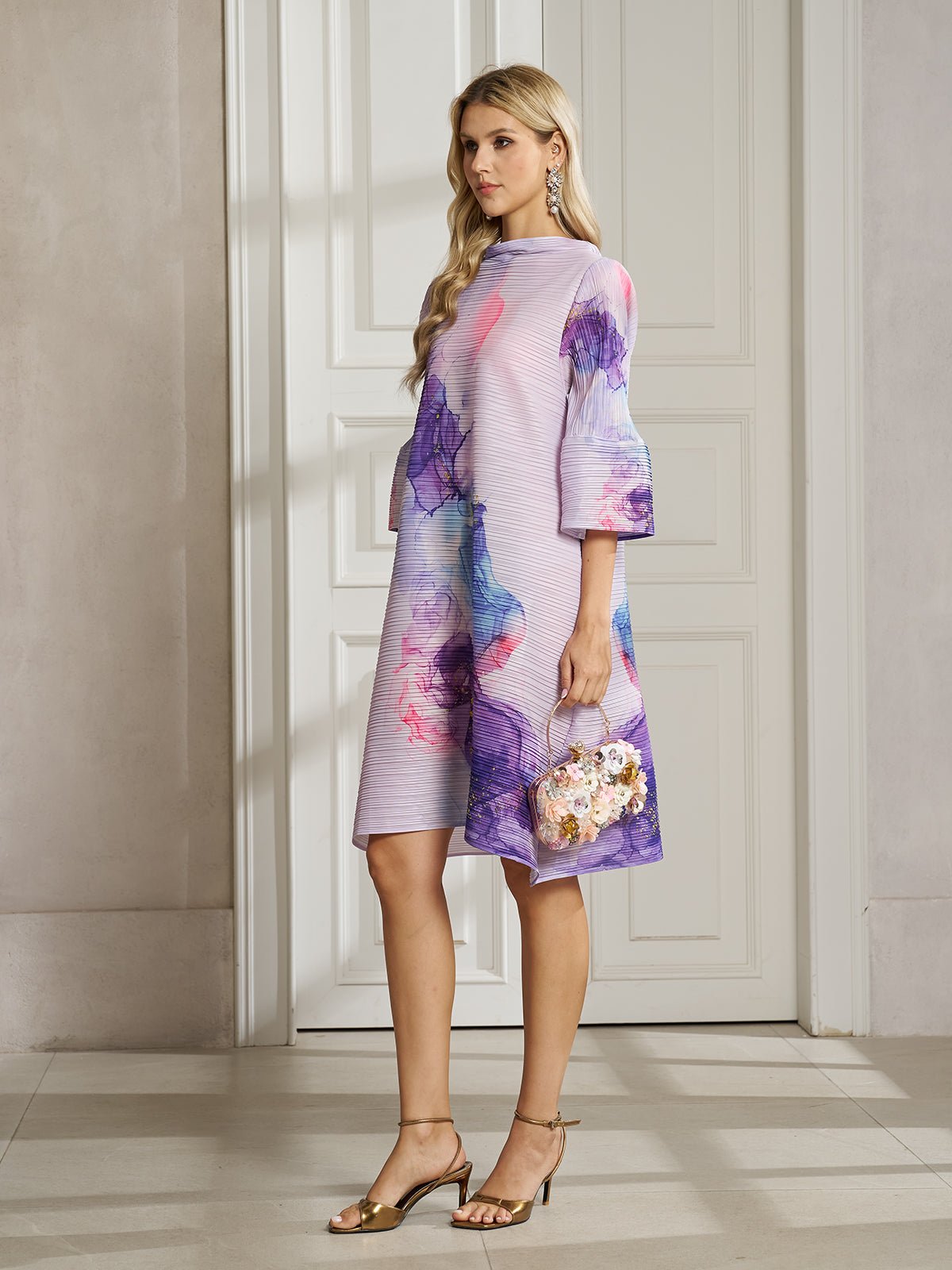 A woman stands indoors in the Social Queen Watercolor Print Plissé Dress, a knee-length bohemian style with flared sleeves crafted from breathable fabric, paired with a floral handbag and gold sandals.