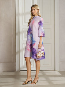 A woman stands indoors in the Social Queen Watercolor Print Plissé Dress, a knee-length bohemian style with flared sleeves crafted from breathable fabric, paired with a floral handbag and gold sandals.