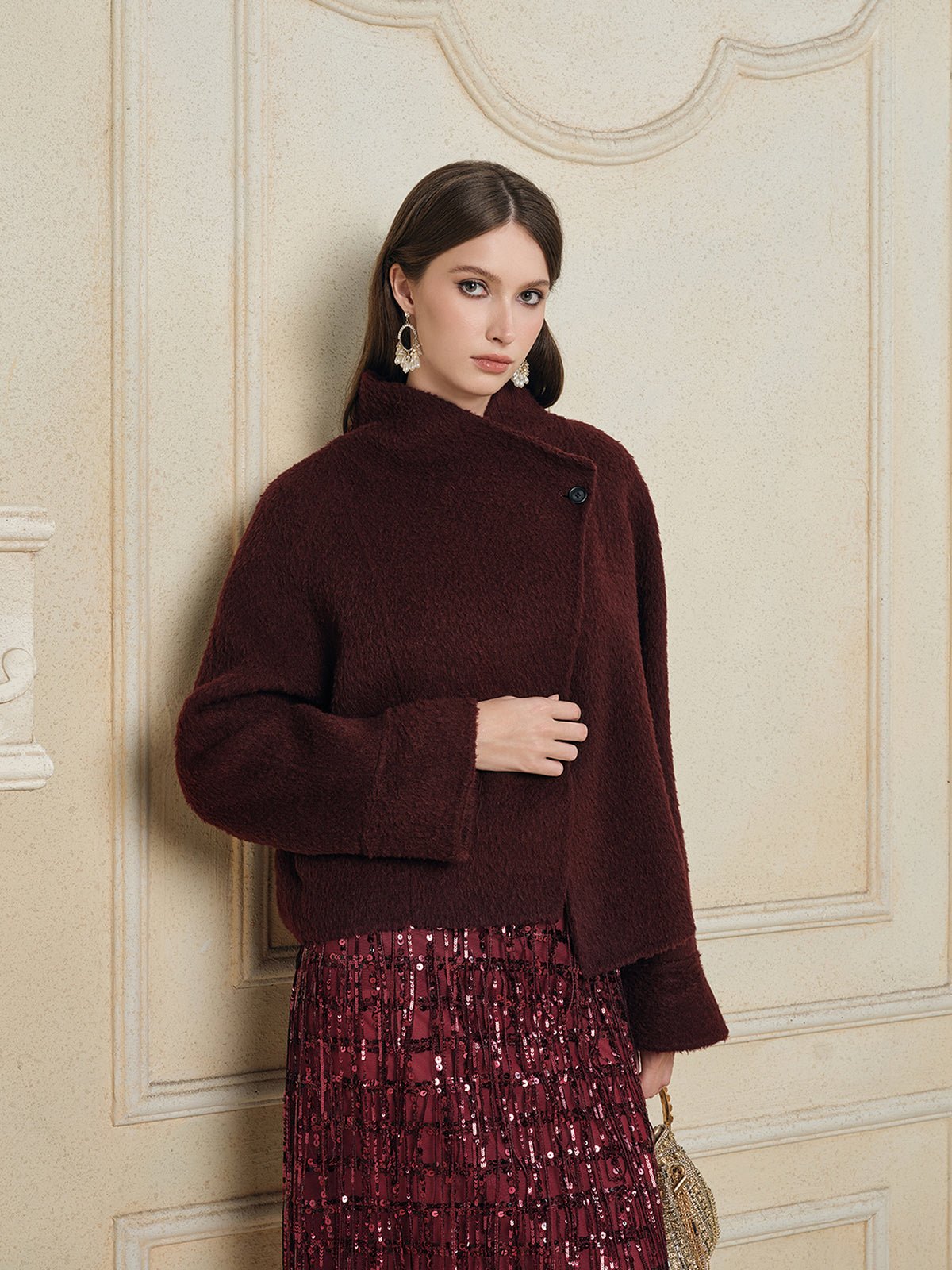 A woman models the Social Queen Wool-Cashmere Cropped Wrap Jacket in burgundy with a sequined skirt, standing before a cream-colored ornate wall.