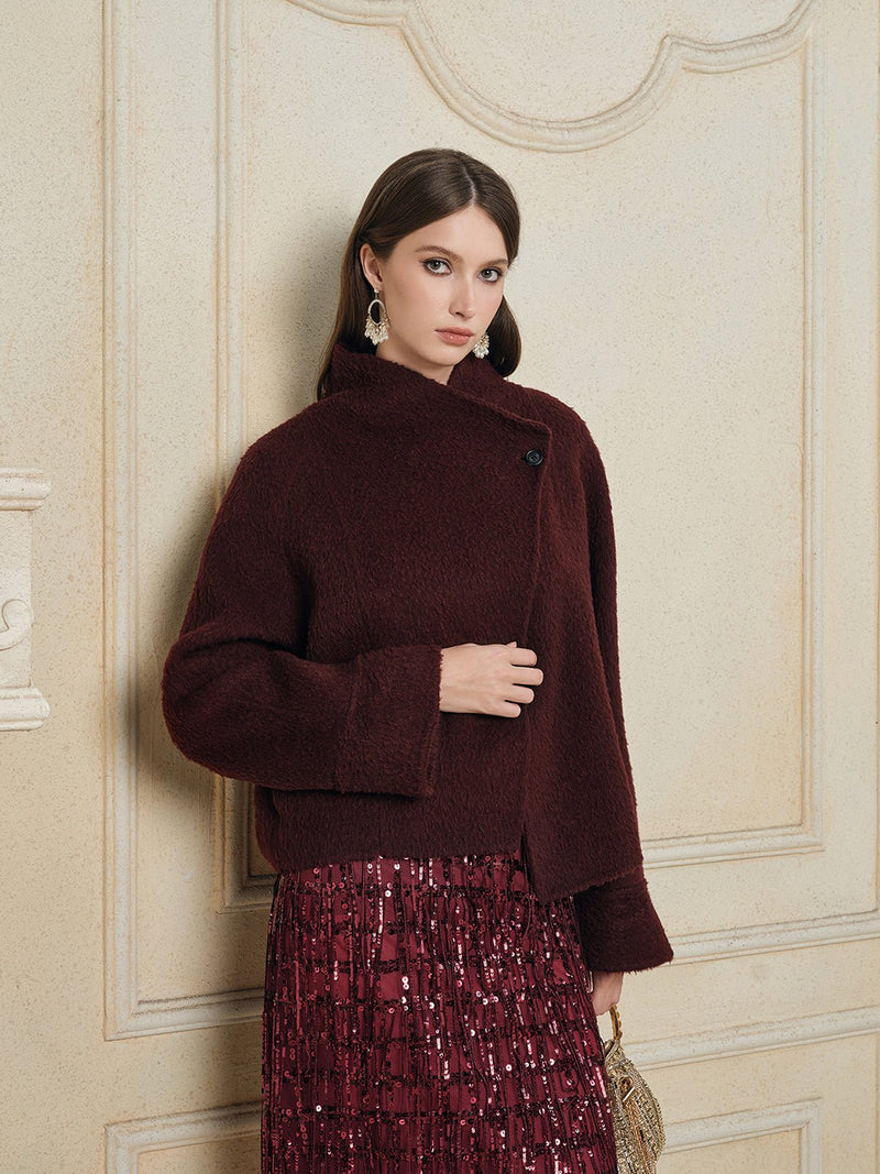 A woman models the Social Queen Wool-Cashmere Cropped Wrap Jacket in burgundy with a sequined skirt, standing before a cream-colored ornate wall.