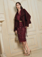 A woman in Social Queen’s Wool-Cashmere Cropped Wrap Jacket stands against an ornate cream wall, wearing a sequined fringed skirt, gold heels, and holding a small gold handbag.