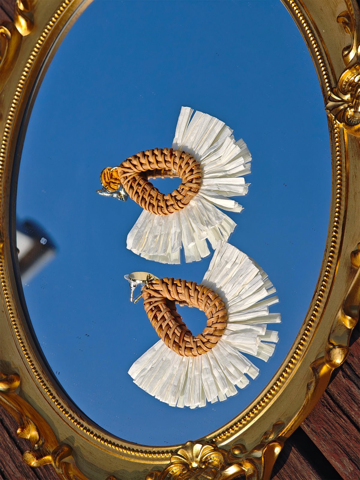 Social Queen’s Woven Fan-Shaped Earrings feature brown, hand-embroidered openwork bases with white fringed edges, elegantly displayed on an ornate gold mirror reflecting the blue sky.