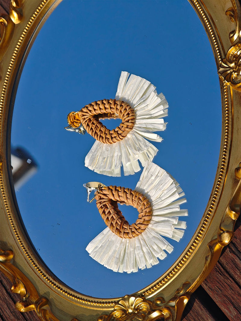 Social Queen’s Woven Fan-Shaped Earrings showcase brown teardrop centers and white fringe, artfully displayed on a gold ornate mirror reflecting the blue sky.