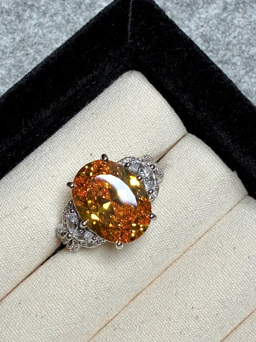 The Social Queen Yellow Crystal Ring features a gold-orange oval crystal with diamond accents on an adjustable white gold band, presented in a black and cream jewelry box—a striking statement piece.